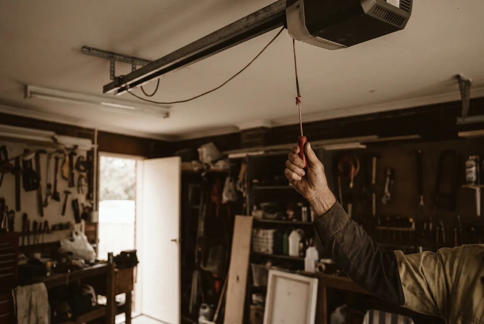 A hand reaching up to pull the red manual release cord hanging from a garage door opener rail